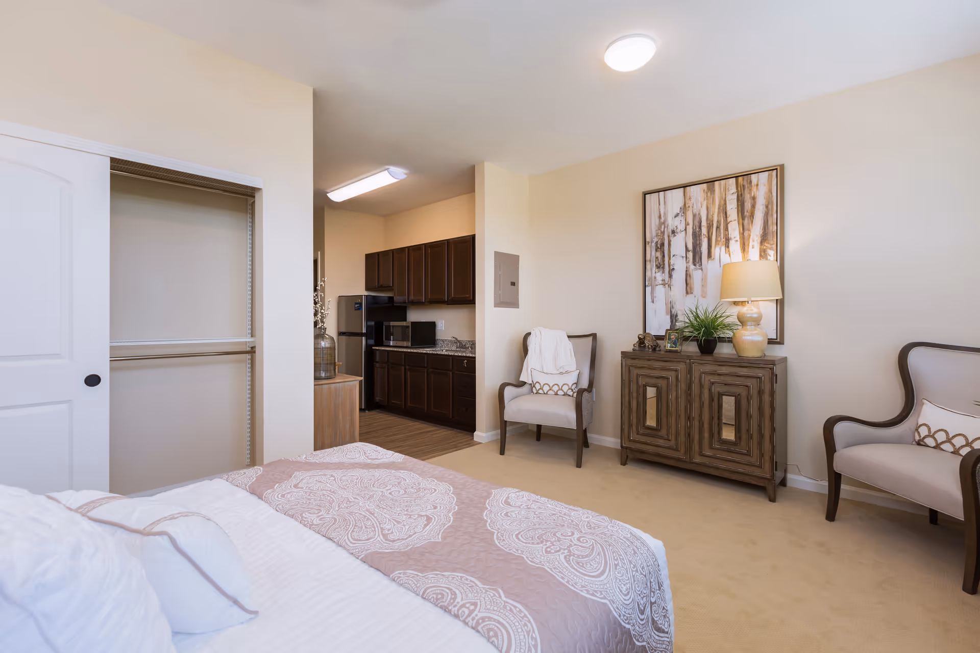 A cozy senior living bedroom featuring a bed with patterned bedding, an open closet with a sliding door, two armchairs with decorative pillows, a wooden cabinet with a lamp and plant, and a kitchenette with dark cabinets, a microwave, and a refrigerator in the background.