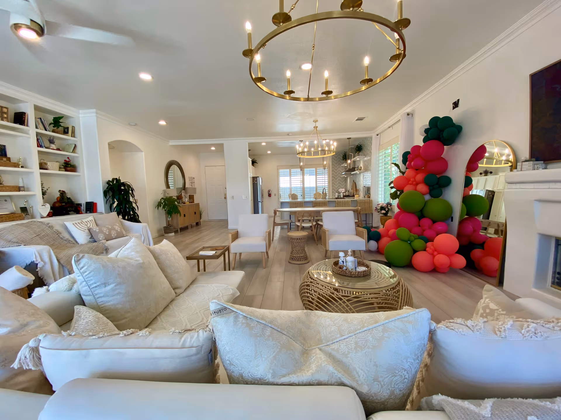 Bright open living room with white sofas, modern chandeliers, and a large colorful balloon display near the fireplace leading into a dining area and kitchen.