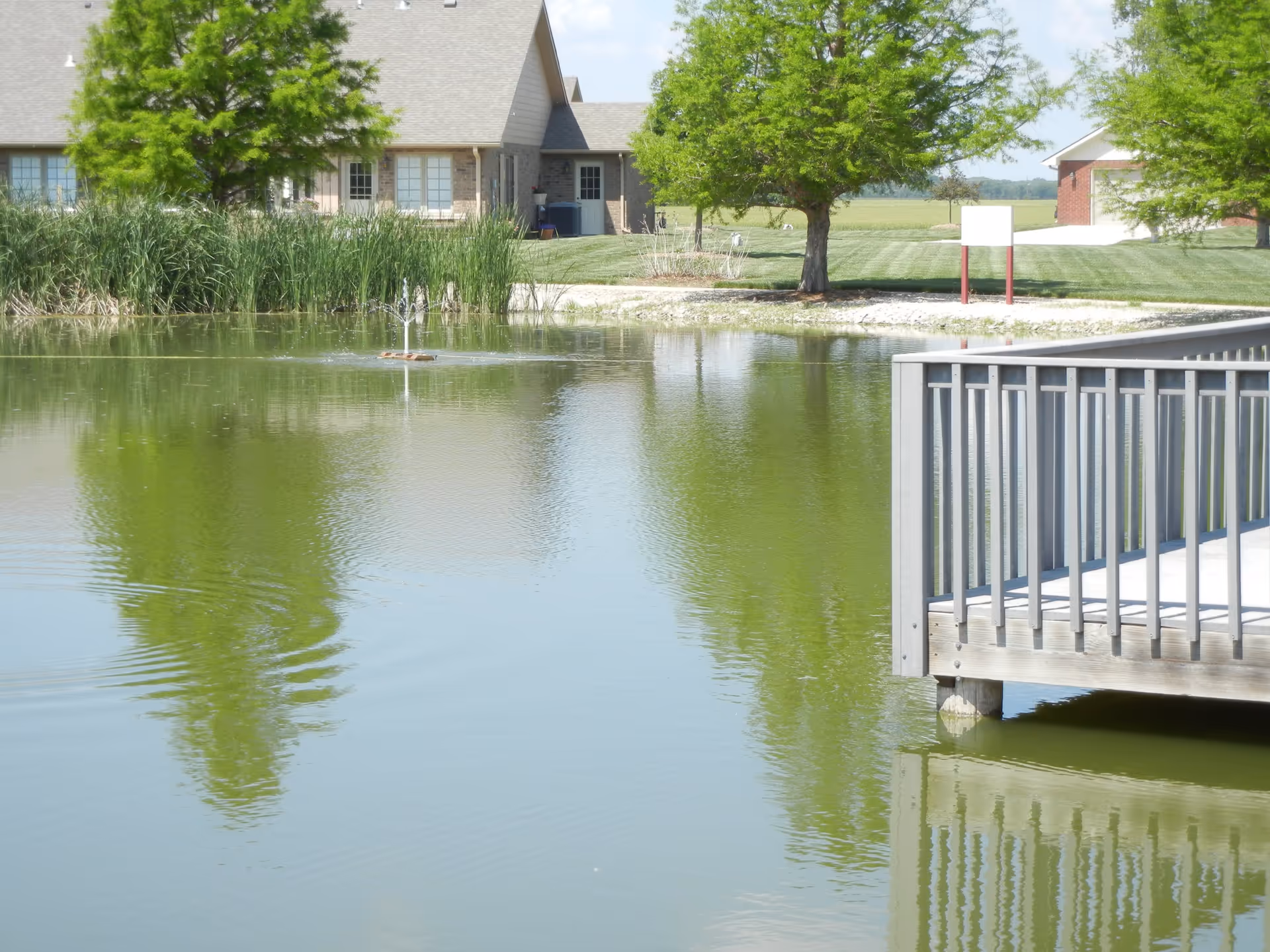 A peaceful outdoor scene featuring a small pond with a water fountain in the center, surrounded by green grass, trees, and residential buildings in the background. A wooden deck with railings extends over the pond on the right side of the image.
