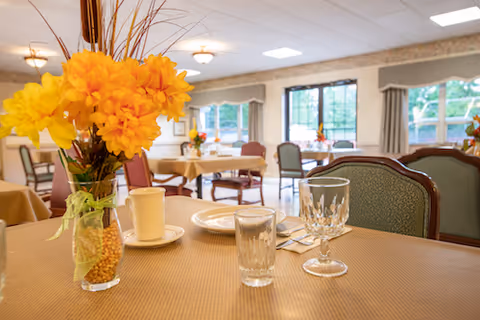 A dining room in a senior living facility with tables covered in beige tablecloths. One table in the foreground has a vase with bright yellow flowers, a white cup and saucer, a plate, a glass, and a goblet. The room has large windows with beige curtains, and several other tables with chairs are visible in the background.