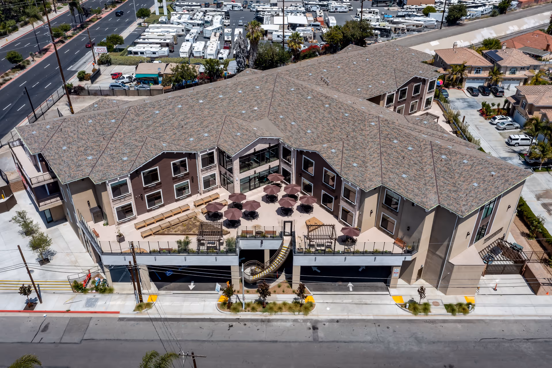 Aerial view of a multi-story senior living facility named Beach Terrace with a large outdoor patio area featuring tables with umbrellas and benches. The building has a brown and beige exterior with a tiled roof. There is a parking garage entrance at the front and surrounding streets and neighboring buildings are visible.