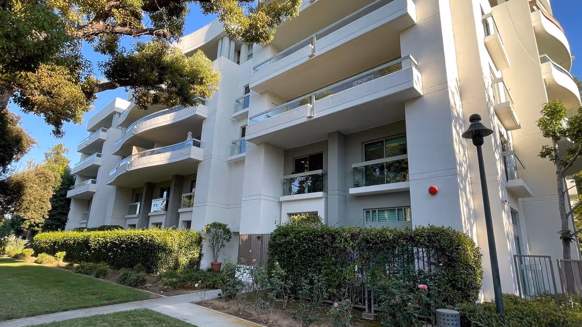 Exterior front view of a multi-story light-colored residential building with balconies, hedges and a sidewalk.