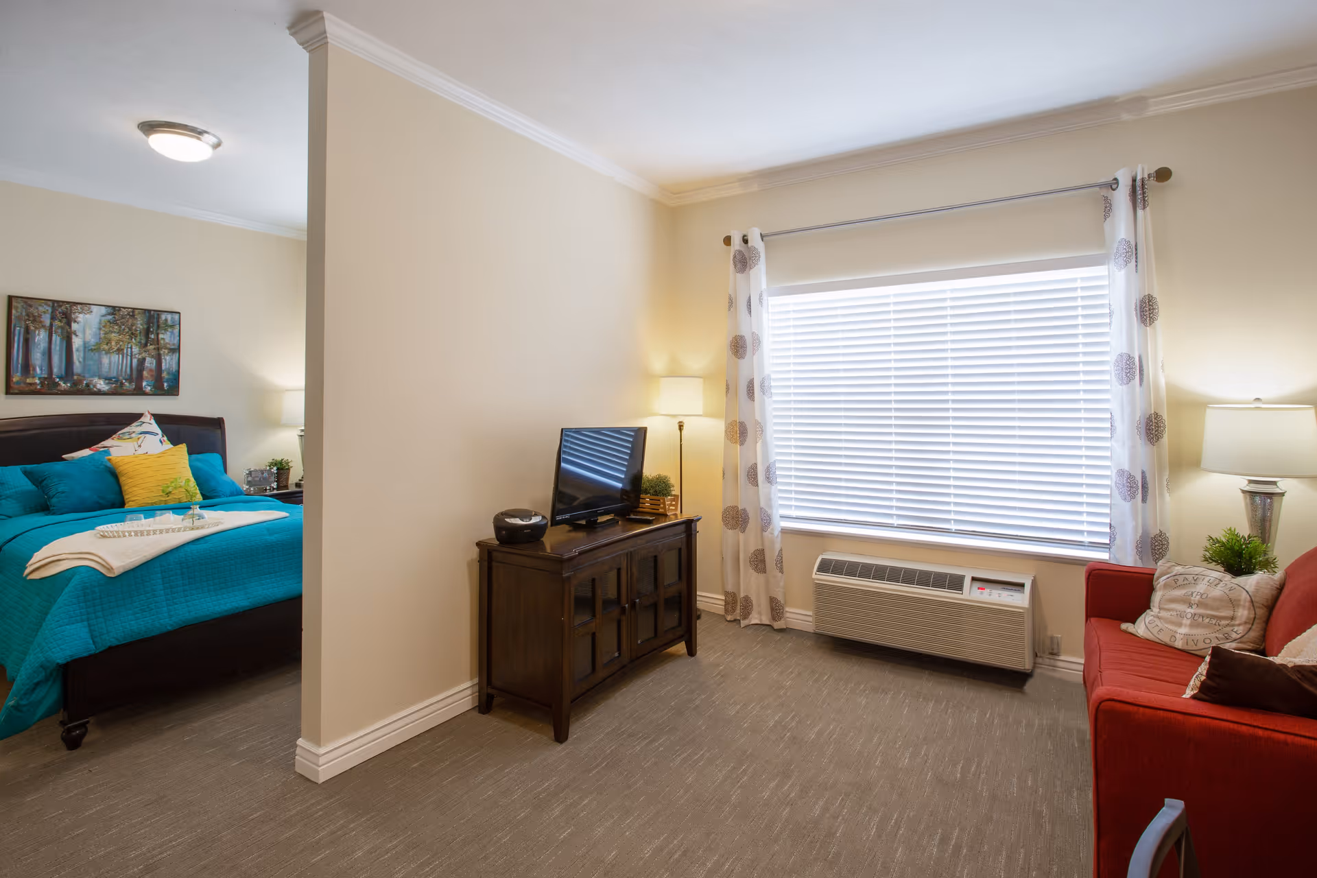 A cozy living area with a red sofa, two lamps, a window with white blinds and patterned curtains, and a wooden TV stand with a flat-screen TV. Adjacent to the living area is a bedroom with a bed covered in teal bedding and colorful pillows, a nightstand with a lamp, and a framed picture on the wall.