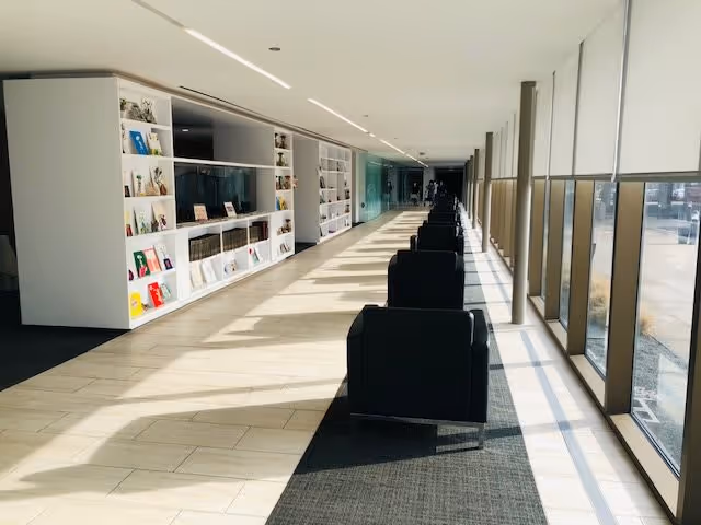 A long, bright hallway with large floor-to-ceiling windows on the right side letting in natural light. On the left side, there is a white built-in bookshelf filled with books and decorative items. A row of black armchairs is aligned along the center of the hallway on a dark carpet runner.