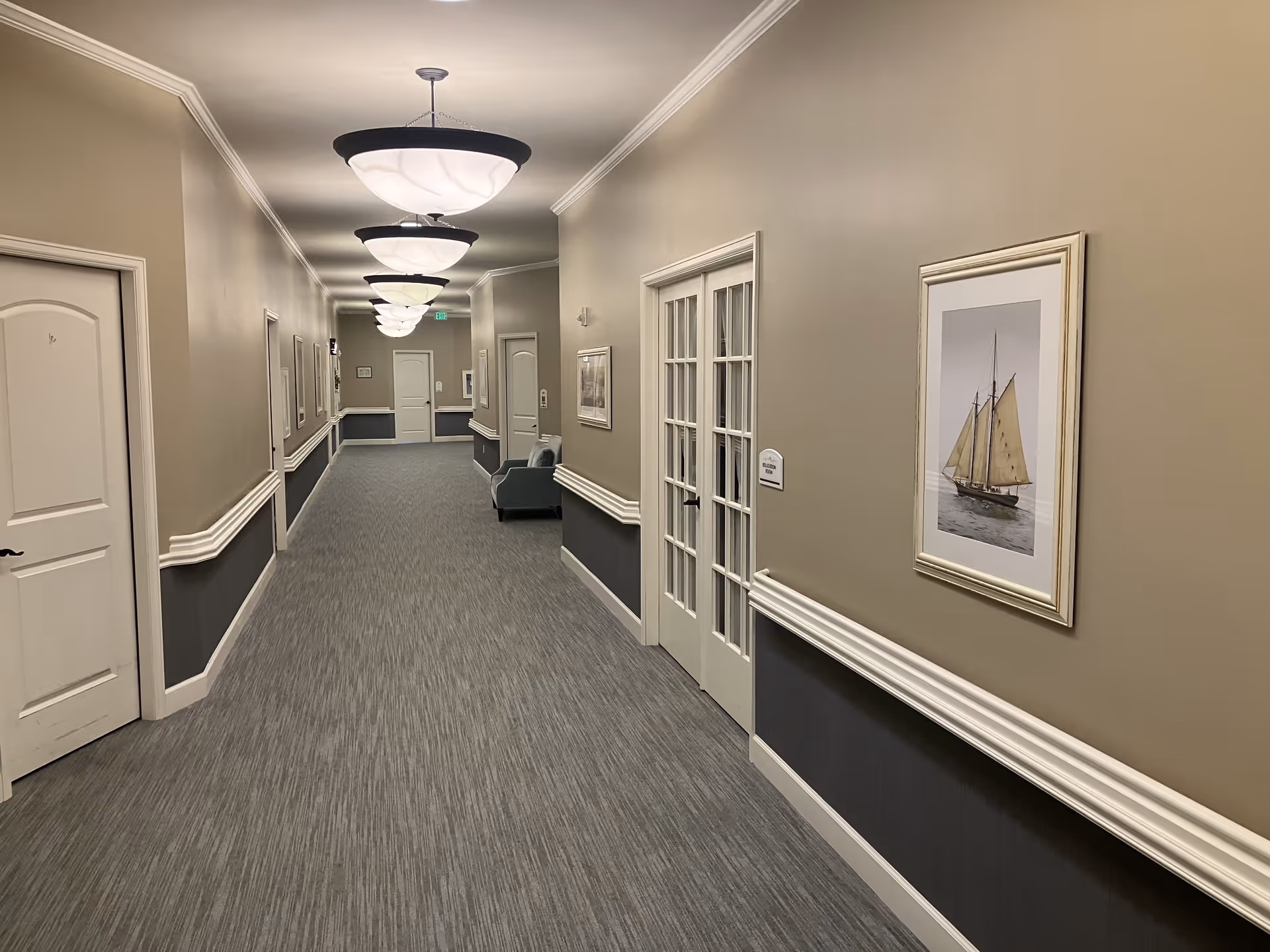A long, well-lit hallway in a senior living facility with beige walls and gray carpet. The walls have white trim and several framed pictures, including one of a sailboat. There are multiple closed doors along the hallway and a pair of French doors on the right side. Ceiling lights with frosted glass shades are evenly spaced along the ceiling. A single armchair is placed against the wall near the French doors.