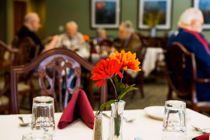 A dining table set with a red folded napkin, two upside-down glasses, a salt shaker, and a small vase holding bright red and orange flowers. In the background, elderly people are seated and engaged in conversation in a dining room setting.