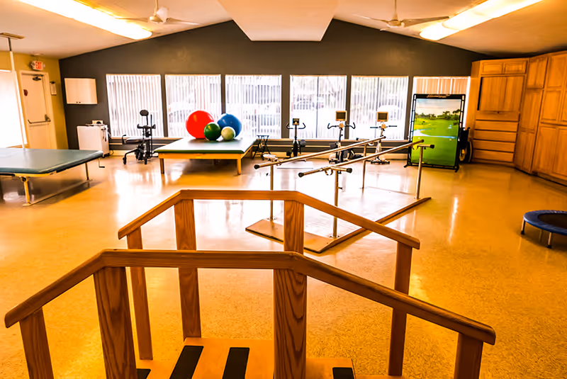 A spacious physical therapy or rehabilitation room with wooden parallel bars for walking exercises in the foreground, exercise tables, colorful therapy balls, stationary bikes, and a small trampoline. Large windows with vertical blinds allow natural light to fill the room.