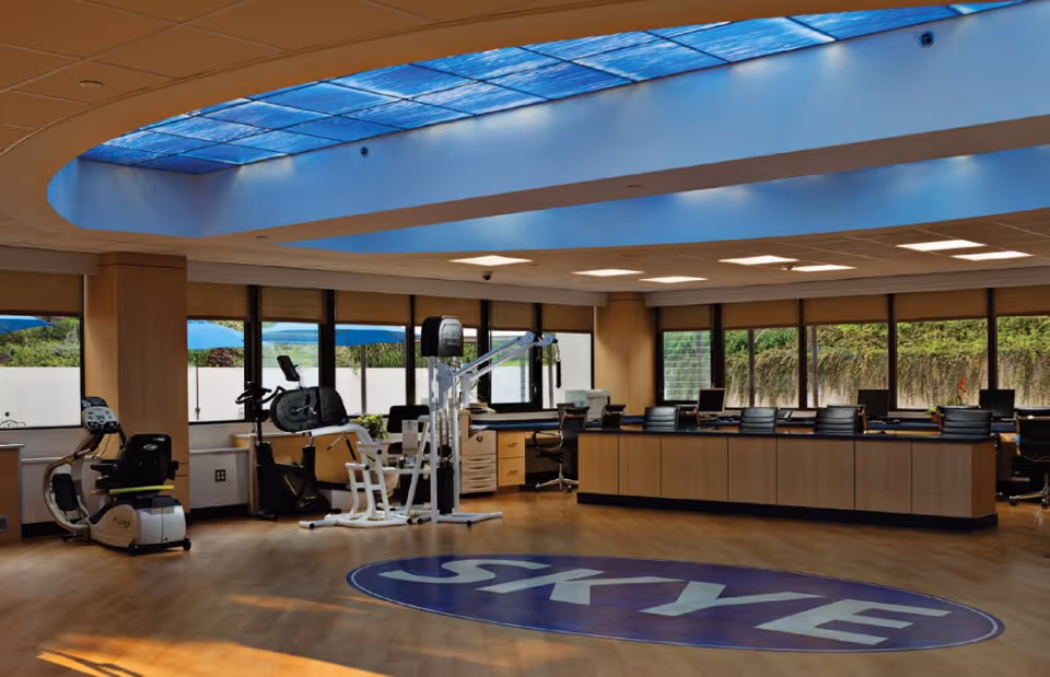 A spacious room with exercise equipment including stationary bikes and a multi-use gym machine, surrounded by large windows letting in natural light. There is a large skylight on the ceiling and a long counter with office chairs and computer monitors along one side. The floor has a large circular logo with the word 'SKYE'.