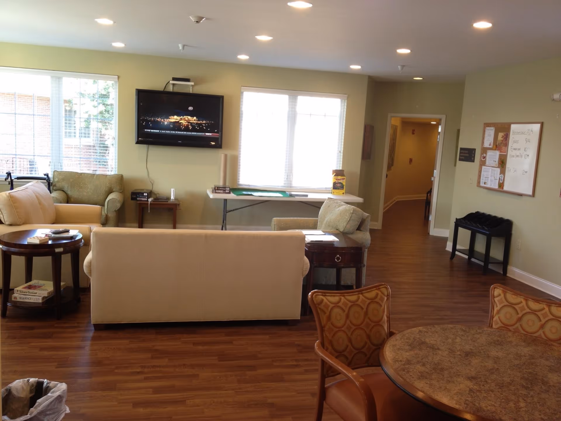 A cozy living room area in an assisted living facility with beige and green sofas, a round table with patterned chairs, a wall-mounted flat screen TV, large windows with blinds, a bulletin board on the wall, and a hallway leading to other rooms. The floor is wooden and the room is well-lit with recessed ceiling lights.