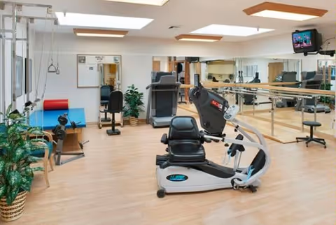 A bright therapy/exercise room with a recumbent exercise bike, treadmill, parallel bars, therapy table, mirrors and a wall-mounted TV.