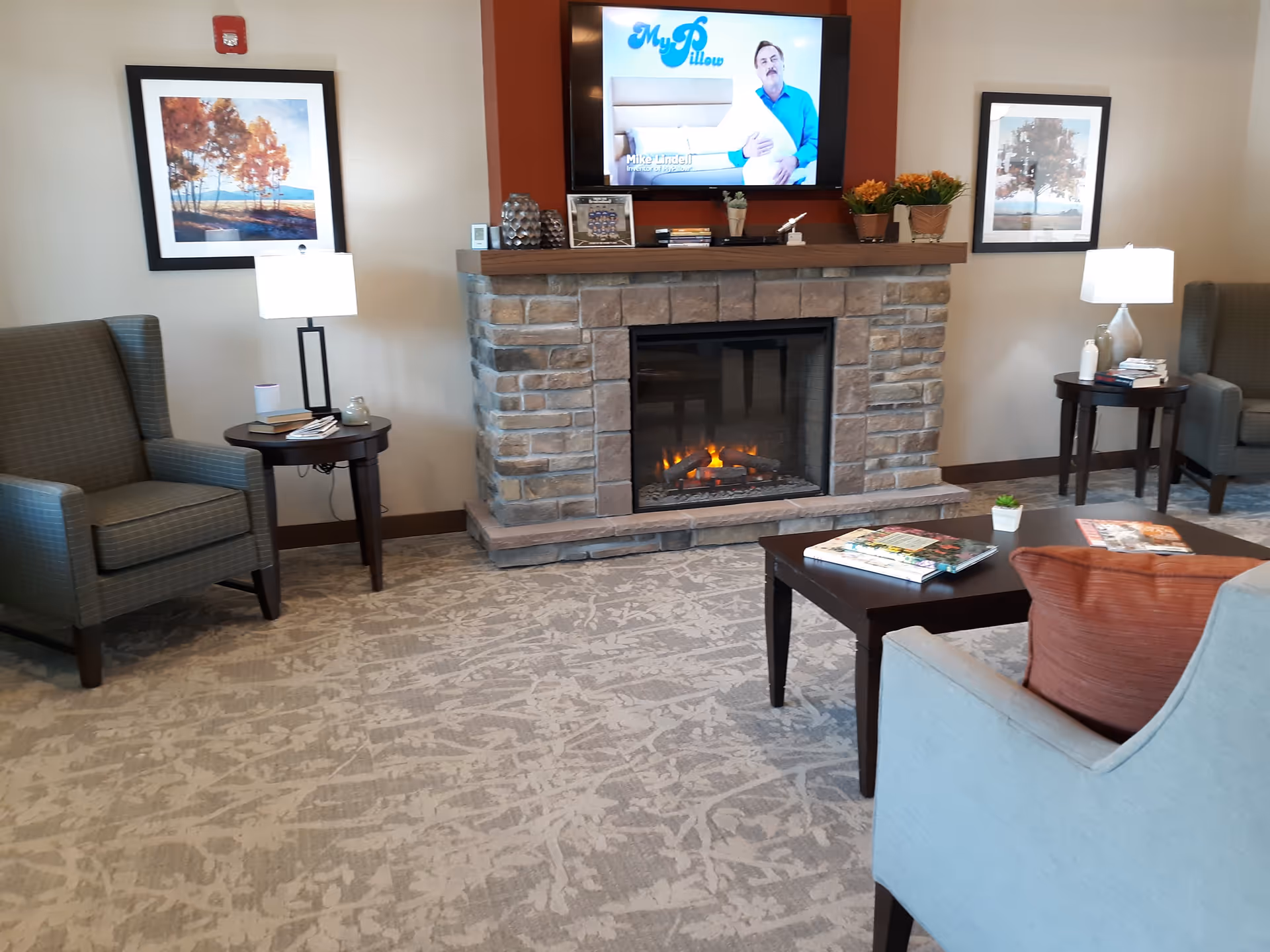 A cozy living room area with a stone fireplace and a TV mounted above it displaying an advertisement for MyPillow. The room features two armchairs with side tables and lamps on either side of the fireplace, a coffee table with magazines, and a light blue chair with an orange cushion in the foreground. Two framed pictures of autumn trees hang on the walls.