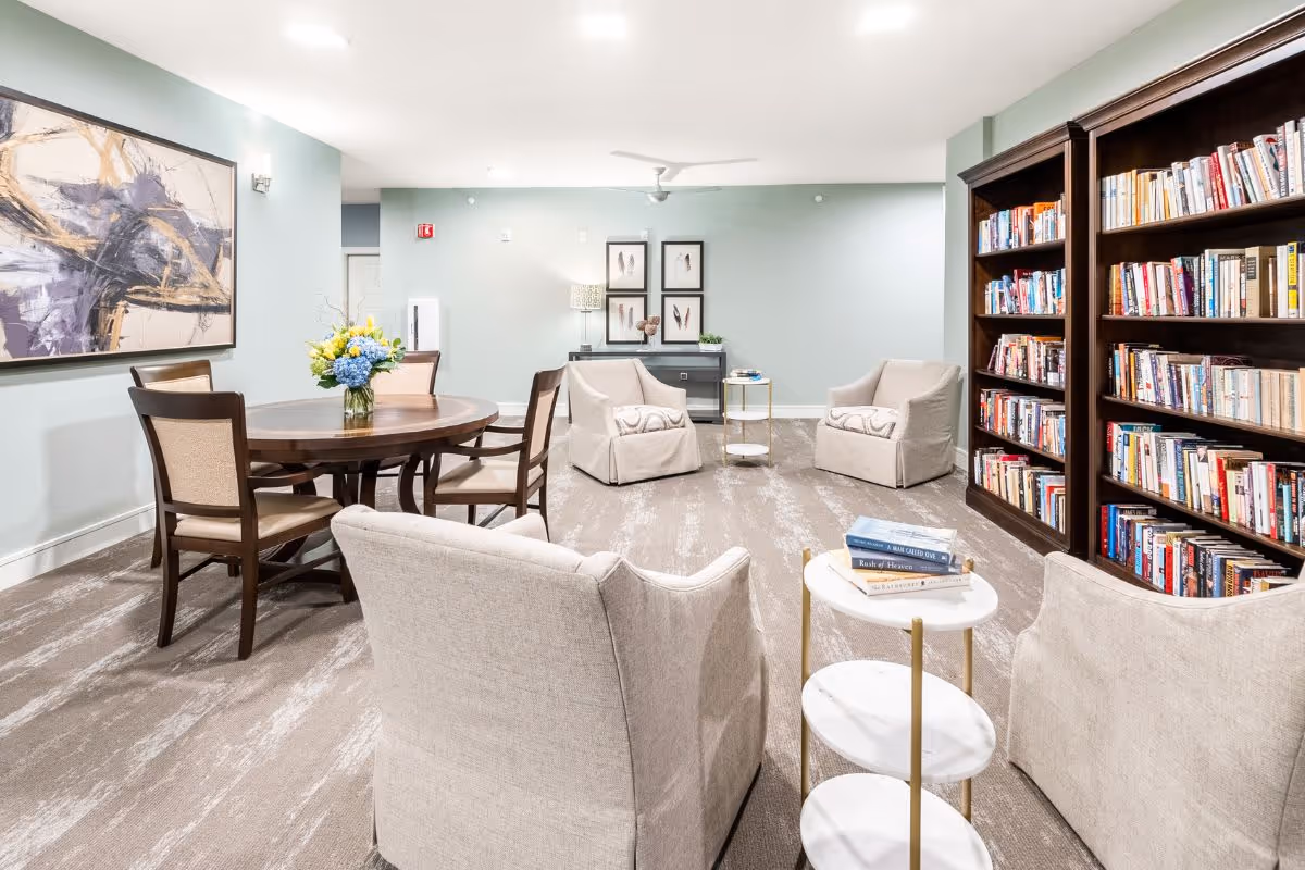 A cozy senior living common area with beige armchairs arranged around small tables, a round wooden table with chairs and a flower vase, and a large bookshelf filled with books against a light blue wall. Abstract artwork and framed feather prints decorate the walls.