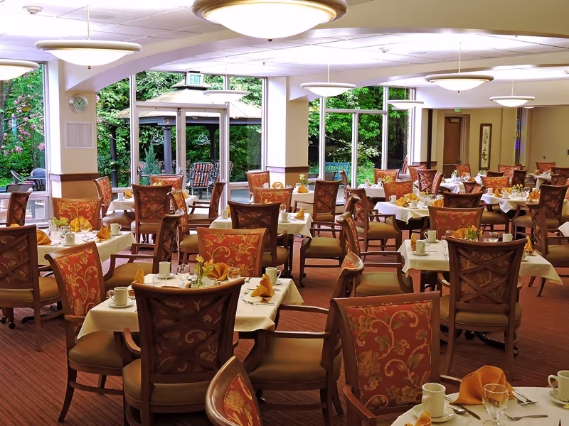 Spacious dining room with many set tables and upholstered patterned chairs, large windows revealing a garden and outdoor seating.