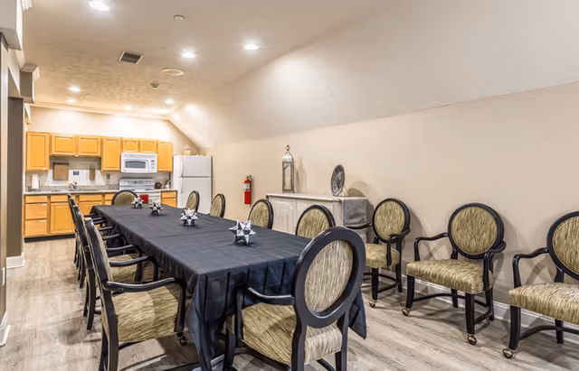 A dining room with a long rectangular table covered with a black tablecloth, surrounded by cushioned chairs with green upholstery and black frames. In the background, there is a kitchen area with wooden cabinets, a white refrigerator, a microwave, and a stove. The room has light-colored walls and wood flooring, with ceiling lights providing illumination.