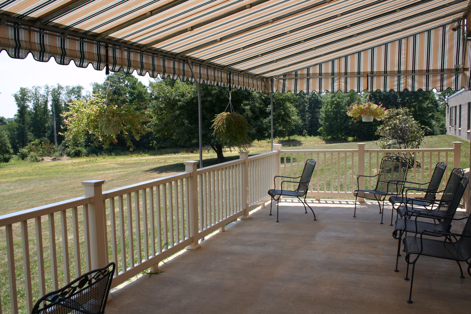 A covered outdoor patio area with a striped awning overhead, several black metal chairs arranged along the railing, and hanging flower baskets. The patio overlooks a grassy area with trees in the background.