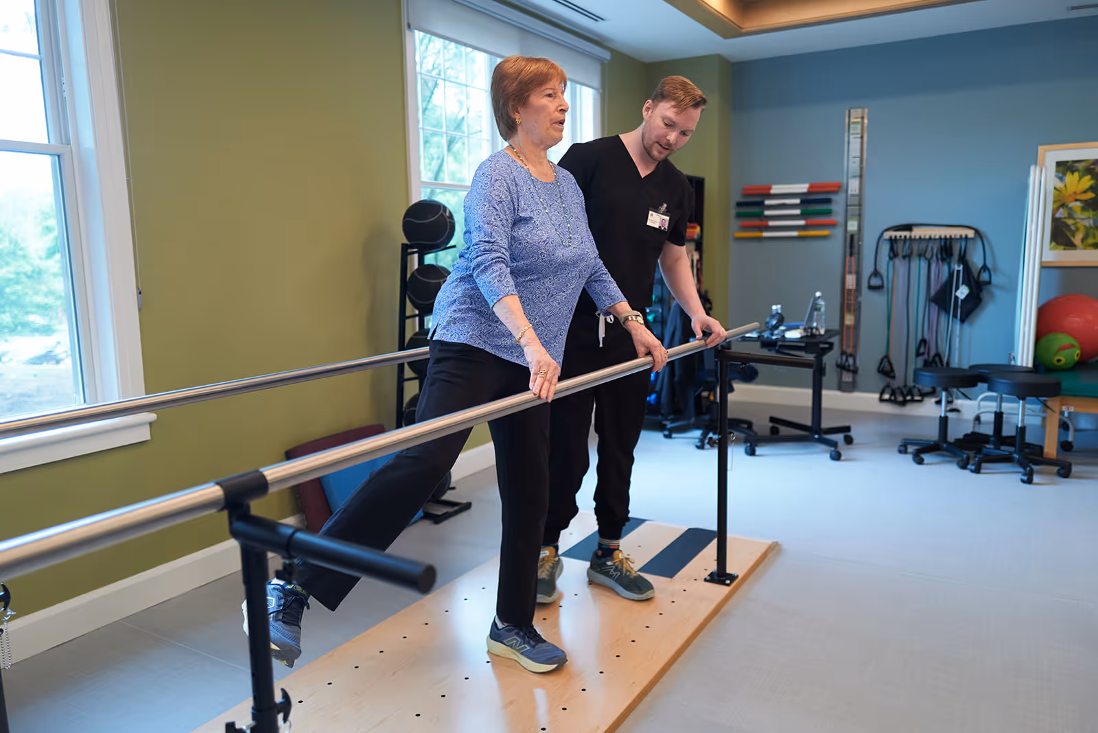 An elderly woman performs a leg exercise using parallel bars in a physical therapy room, assisted by a male therapist. The room has exercise equipment, resistance bands, and a large window letting in natural light.