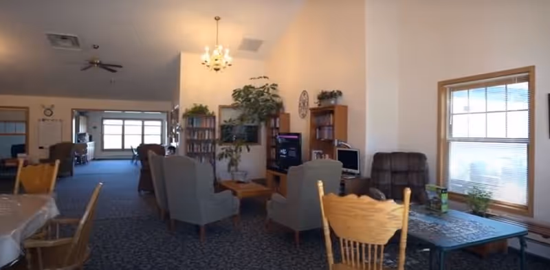 Interior view of a senior living facility common area with multiple seating arrangements including armchairs and wooden chairs around tables. There is a television, bookshelves, and large windows letting in natural light. The room has a carpeted floor and light-colored walls with some plants and decorative items.