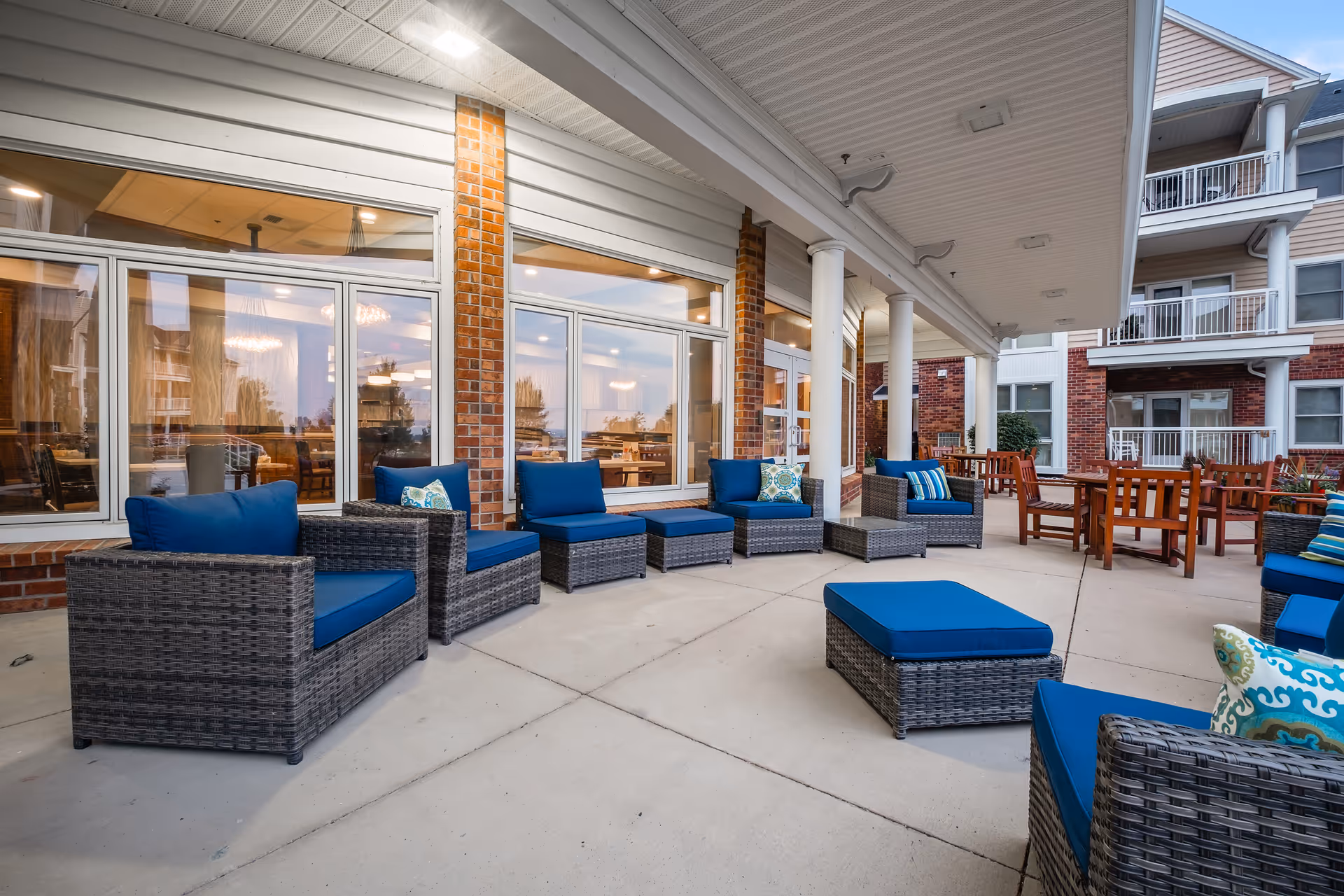 Outdoor patio area at St. John's United Mission Ridge featuring wicker chairs and ottomans with blue cushions arranged on a concrete floor under a covered roof. There are large windows and brick pillars along the building wall, and wooden tables and chairs are visible further back on the patio.