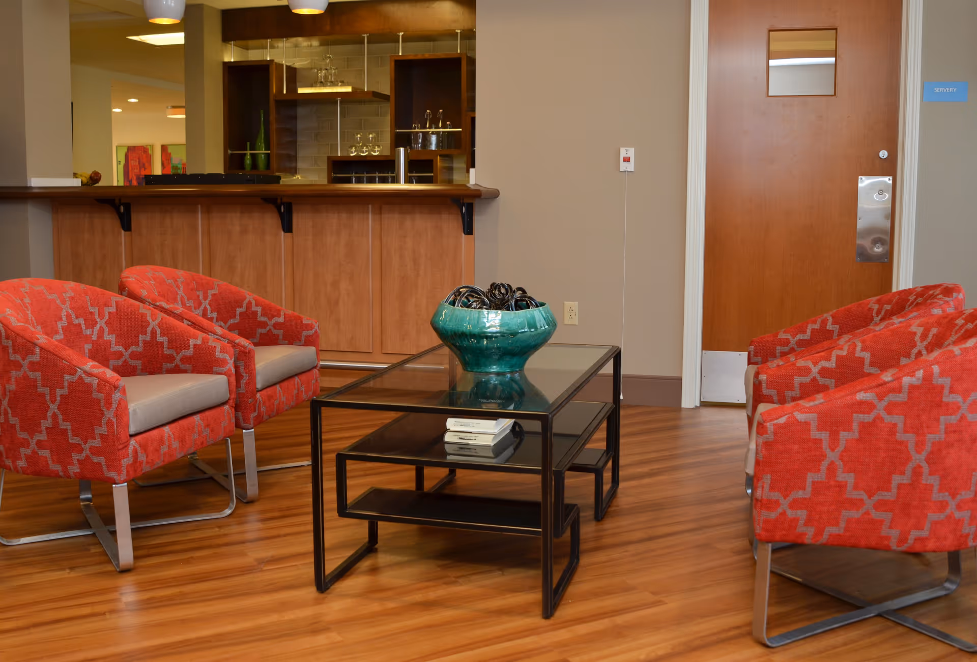 A seating area with four red patterned armchairs around a glass coffee table topped by a large green bowl, with a wooden counter and a door in the background.