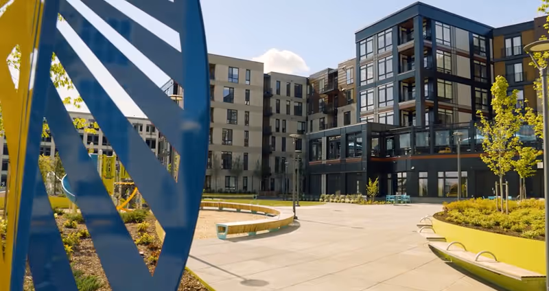 Outdoor courtyard area of a senior living facility with modern multi-story buildings in the background, landscaped garden beds with small trees, curved benches, and a blue and yellow decorative structure in the foreground.