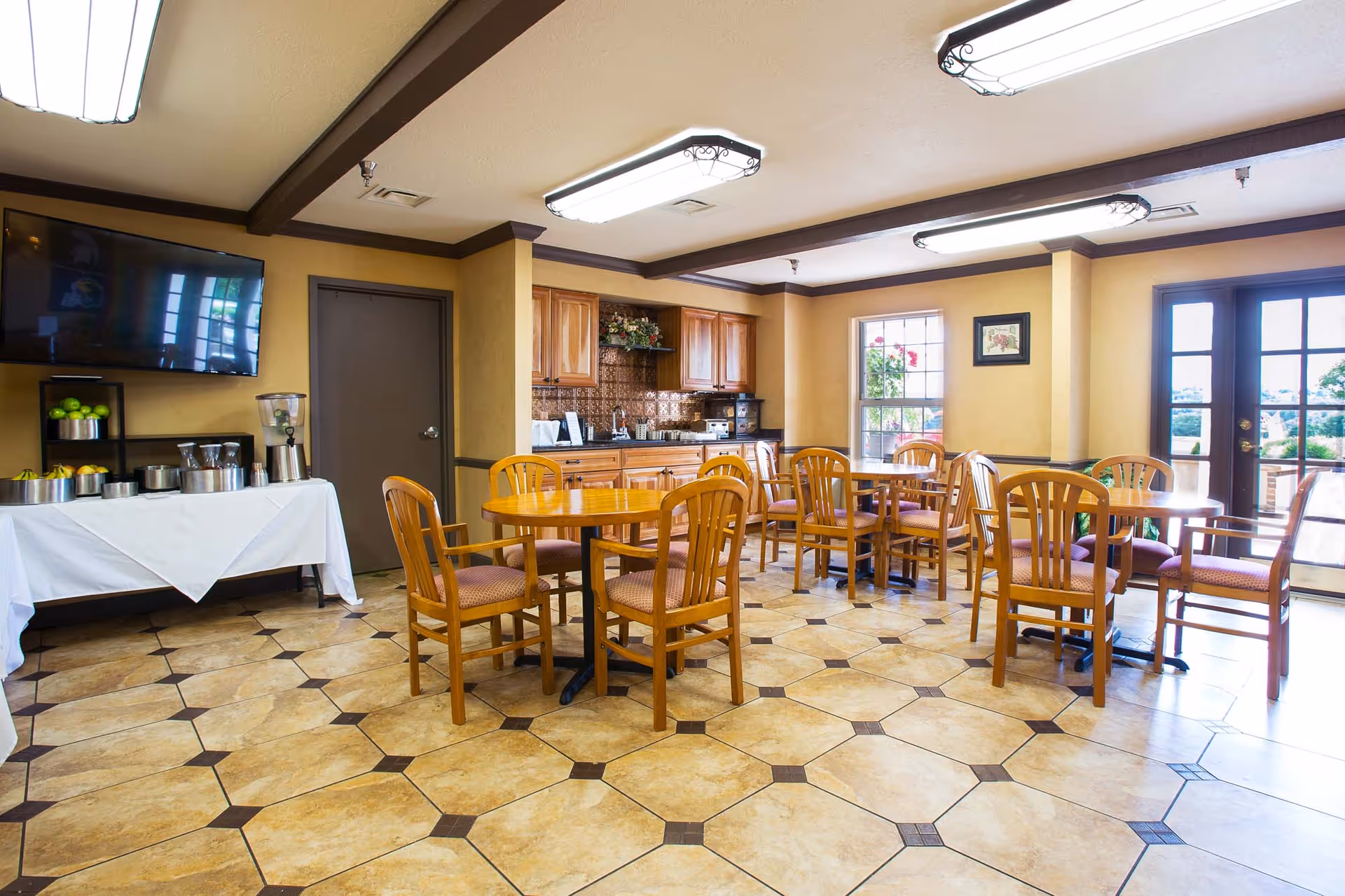 A bright dining area with several wooden tables and chairs arranged on a tiled floor. The room has beige walls with dark trim and a kitchenette with wooden cabinets and a countertop in the background. A large flat-screen TV is mounted on the wall above a table with a white tablecloth holding a beverage dispenser and bowls of fruit. There are windows and glass doors letting in natural light.