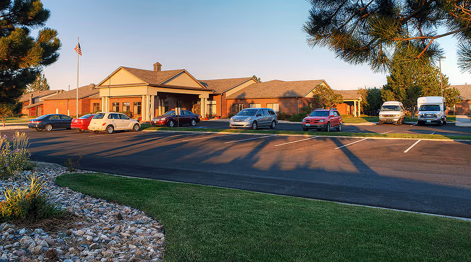 Front exterior of a single-story brick senior living facility with a covered drive-up entrance, parked cars, and an American flag.
