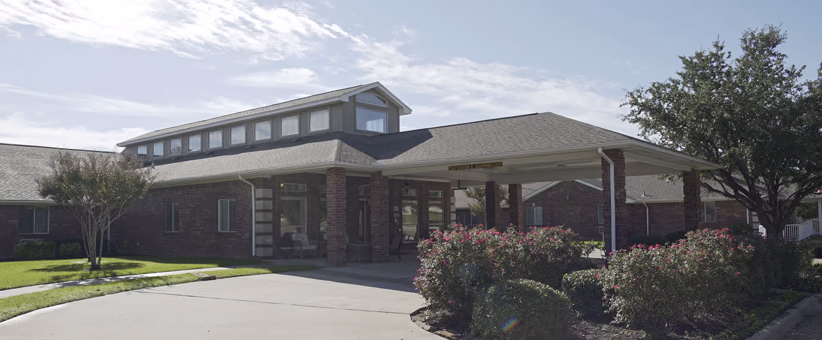 Exterior view of a single-story brick building with a covered entrance, surrounded by green lawns, bushes, and trees under a partly cloudy sky.