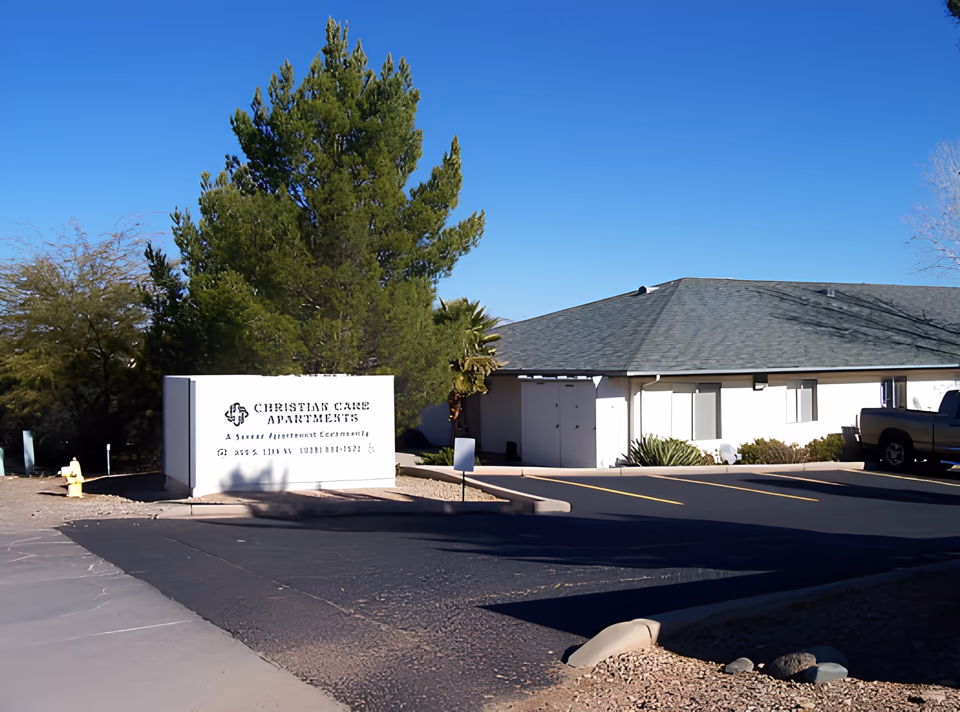 Exterior view of Christian Care Apartments with a white sign displaying the facility name and description. The building has a gray roof and white walls, surrounded by trees and a clear blue sky.