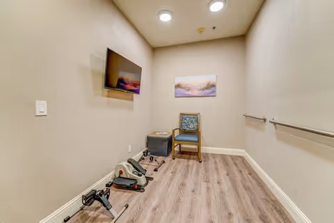 A small exercise room with light wood flooring and beige walls. The room contains a stationary pedal exerciser on the floor, a single chair with a patterned cushion, a small black ottoman, and a wall-mounted flat-screen TV. There is a landscape painting on the wall above the chair and a handrail along one wall. The ceiling has recessed lighting.