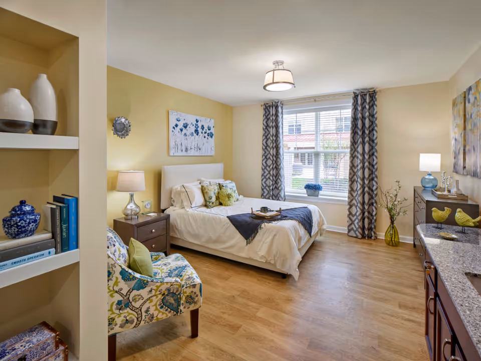 A bright and cozy bedroom with a large window covered by patterned curtains. The room features a neatly made bed with white bedding and decorative pillows, a wooden nightstand with a lamp, a floral upholstered chair with a green cushion, and a dresser with decorative items including two yellow bird figurines and a lamp. The walls are painted a soft yellow, and there is a piece of floral artwork hanging above the bed. The floor is light wood, and there are shelves with books and vases on the left side.