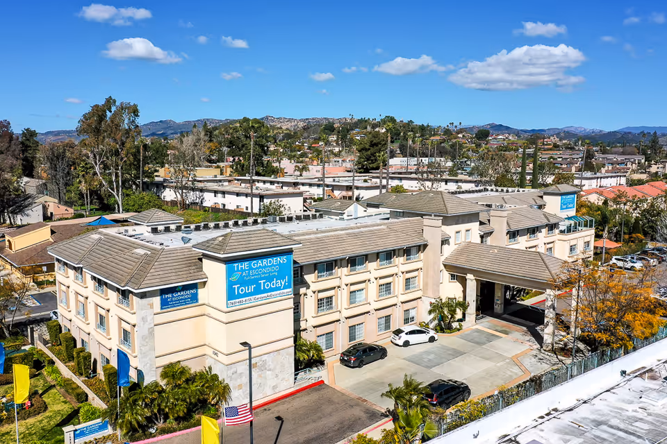 Aerial view of The Gardens at Escondido, a multi-story senior living facility with beige exterior walls and a tiled roof. The building has a covered entrance with a driveway and several parked cars. Surrounding the facility are trees, residential buildings, and hills in the background under a blue sky with scattered clouds.