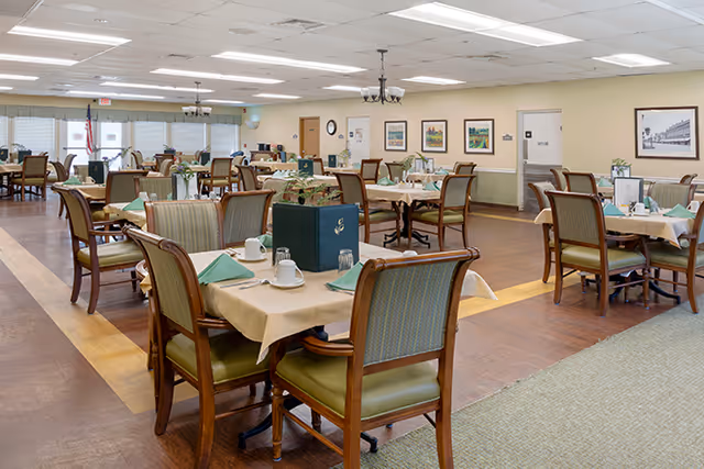 A spacious dining room in a senior living facility with multiple tables set for meals. Each table has beige tablecloths, green napkins, cups, and glasses. The room has wooden chairs with green upholstery, framed artwork on the walls, and ceiling lights. There is an American flag visible in the background near large windows with blinds.