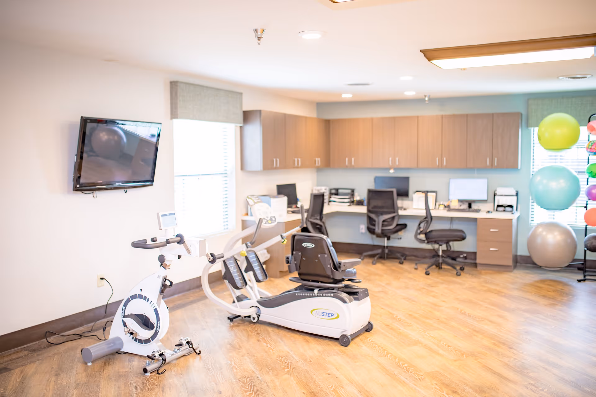 Bright physical therapy room with stationary and recumbent exercise bikes, stability balls, and a workstation area with desks and computers.