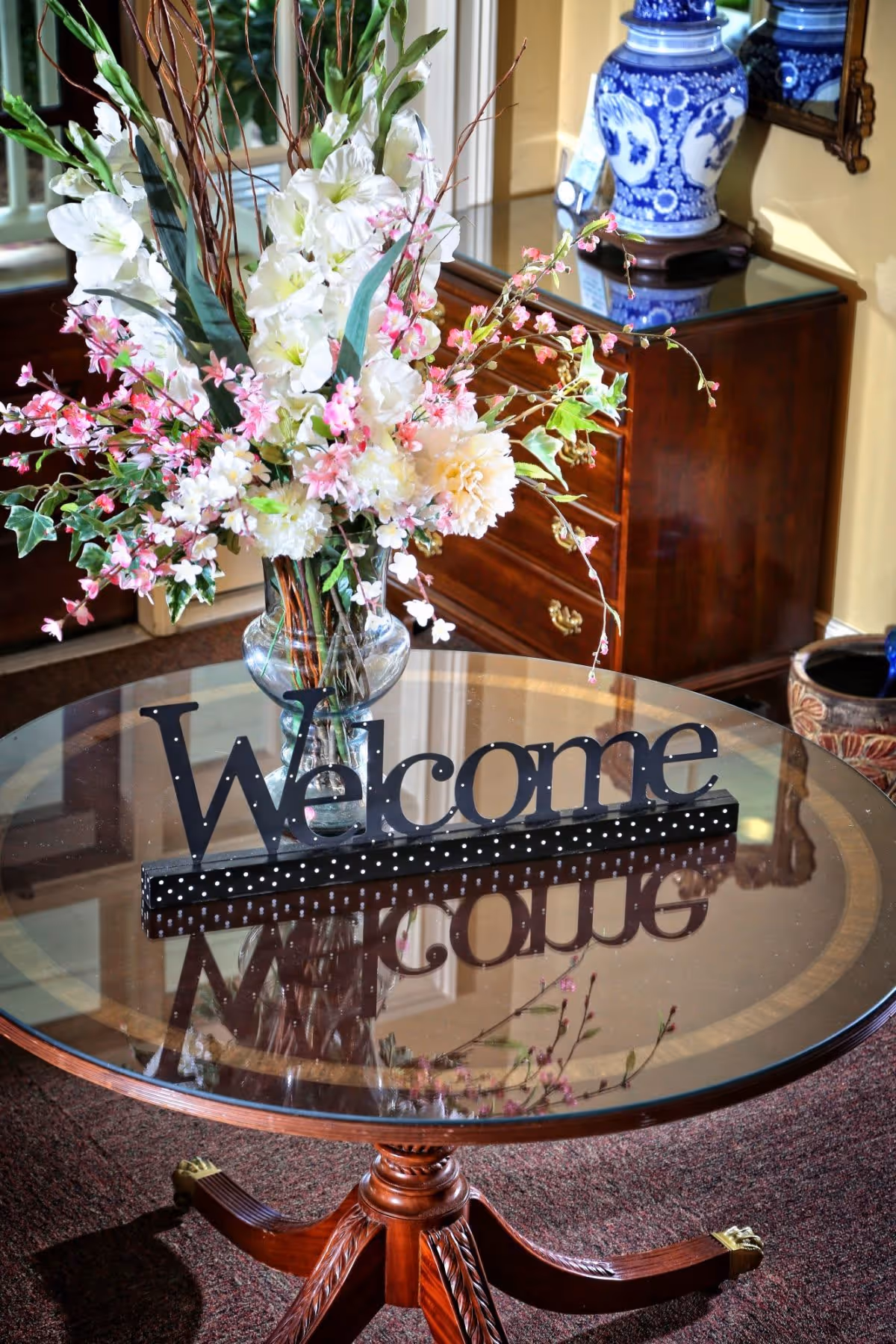 A round glass-top wooden table with a decorative 'Welcome' sign and a large floral arrangement featuring white and pink flowers. In the background, there is a wooden chest of drawers with a blue and white ceramic vase on top and a mirror on the wall.