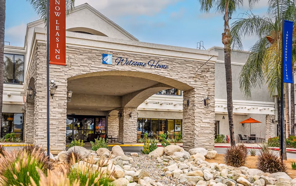 Exterior view of Menifee Senior Living facility entrance with stone pillars and an archway displaying a 'Welcome Home' sign. There are palm trees, landscaping with rocks and plants, and banners on poles reading 'Now Leasing' and 'Independent Living'.