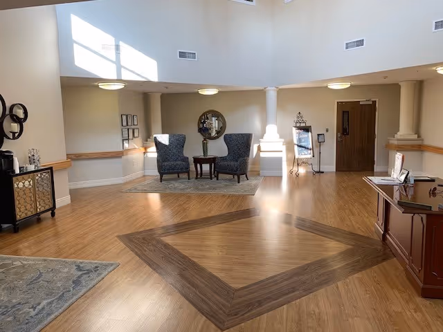 A bright and spacious interior common area with wooden flooring featuring a darker wood diamond pattern in the center. Two upholstered armchairs with a small round table between them are placed on a rug against the far wall, beneath a round mirror. The room has high ceilings with recessed lighting and large windows allowing natural light to flood in. There are decorative elements on the walls and furniture including a cabinet on the left and a reception desk on the right.