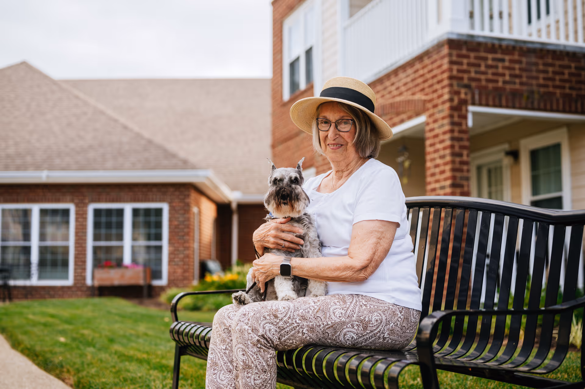 An elderly woman wearing a hat and glasses sits on a black metal bench outside a brick building, holding a small gray dog on her lap. The background shows green grass, windows, and part of the building exterior.