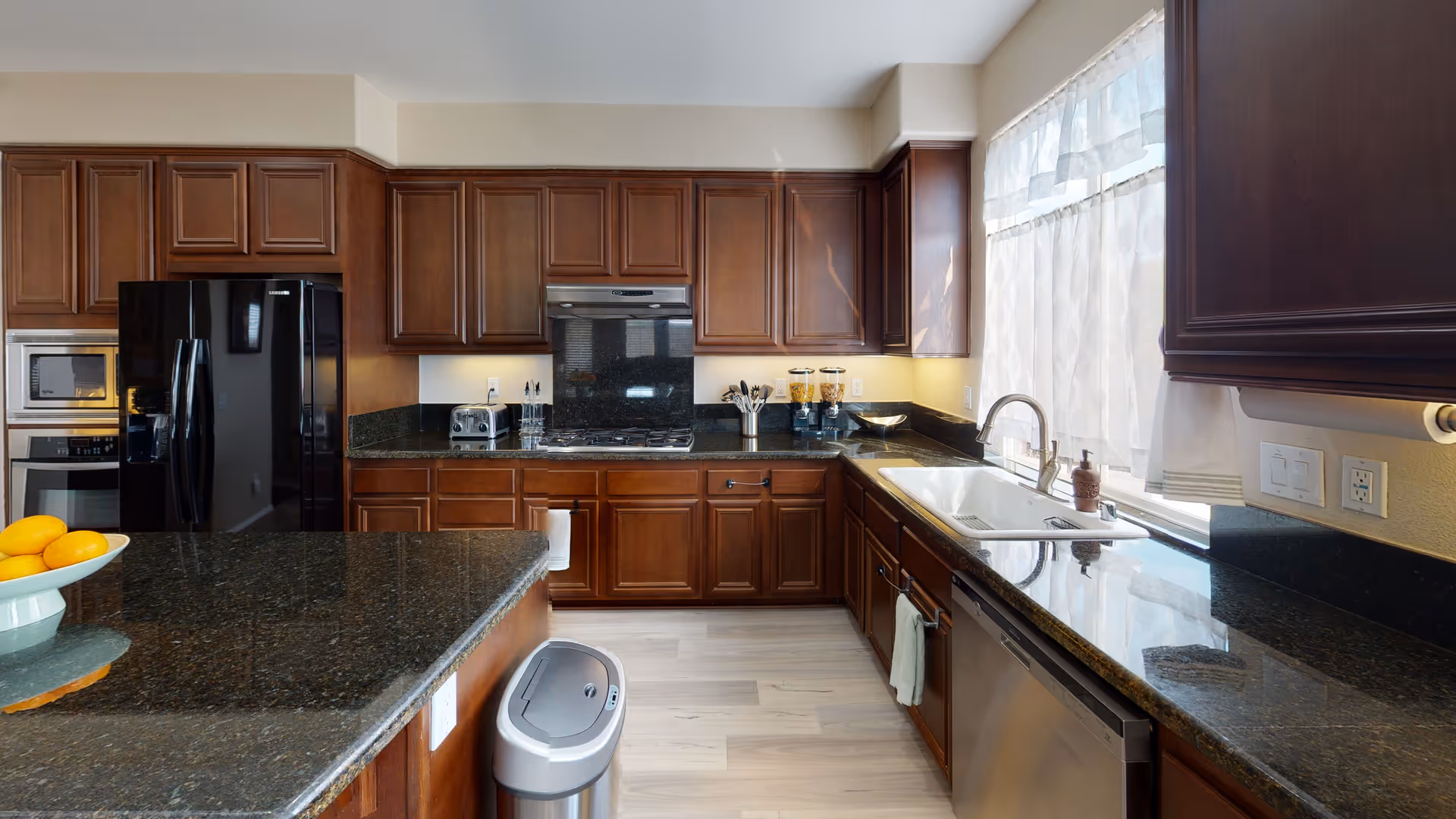 A modern kitchen with dark wooden cabinets, black granite countertops, a black refrigerator, built-in microwave and oven, a stainless steel dishwasher, a white sink under a window with sheer curtains, and a kitchen island with a bowl of oranges. There is also a stainless steel trash can on the floor.