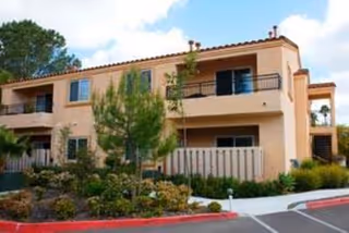 Two-story beige stucco residential building with balconies, landscaped front and a parking curb.