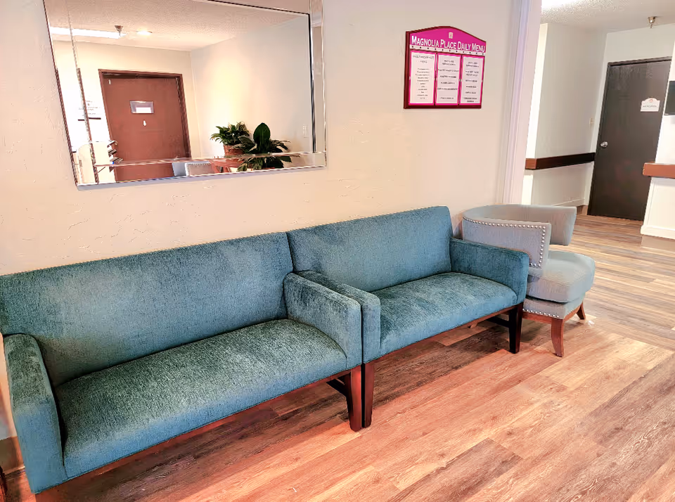 A waiting area with two teal upholstered sofas and a gray armchair on a wooden floor. A large mirror hangs on the wall above the sofas, reflecting a door and a plant. A purple-framed daily menu board for Magnolia Place is mounted on the wall next to the sofas. There are two dark-colored doors visible in the hallway.