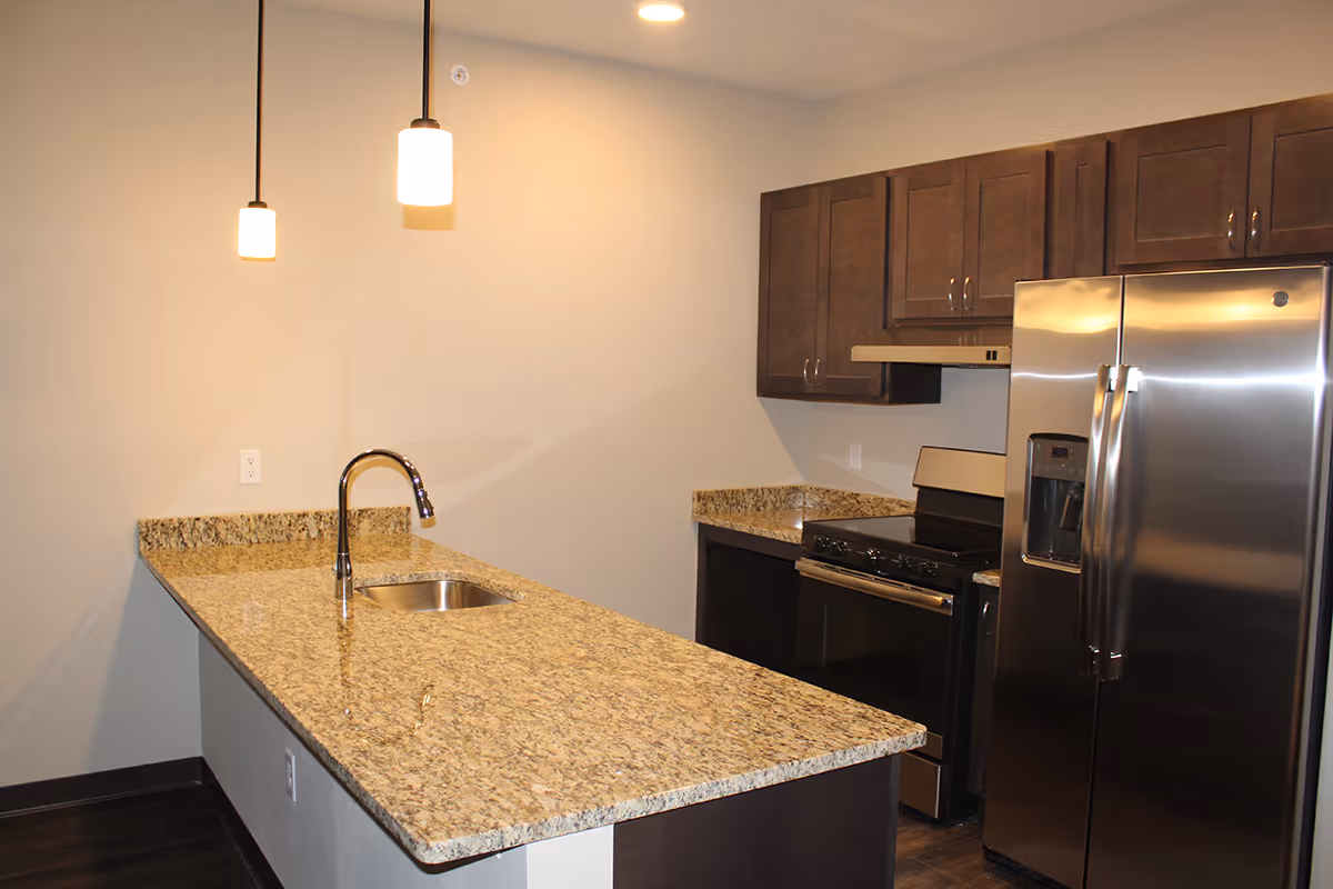 Modern kitchen with granite countertops, a stainless steel refrigerator, stove, and dark wood cabinets. Two pendant lights hang over the countertop with a built-in sink and faucet.