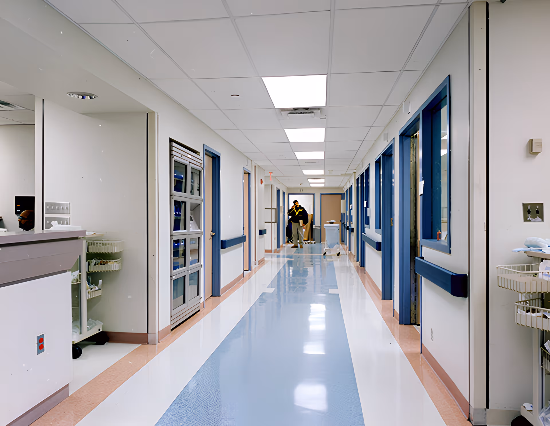 Long, clean interior corridor of a senior care/medical facility with blue-trimmed doors and a person standing at the far end.