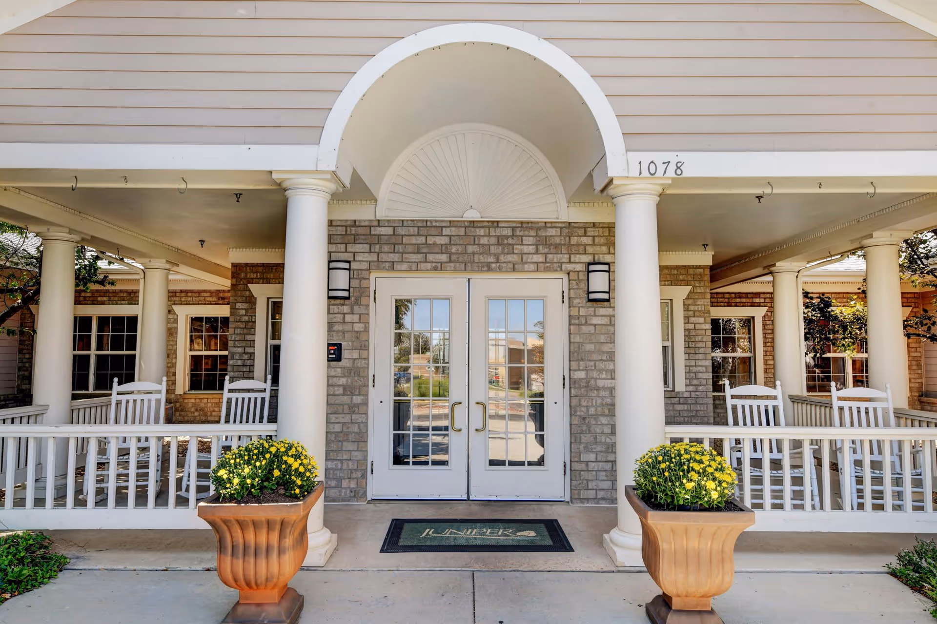 Entrance of a senior living building with double glass doors, white columns, a covered porch with rocking chairs and potted flowers.
