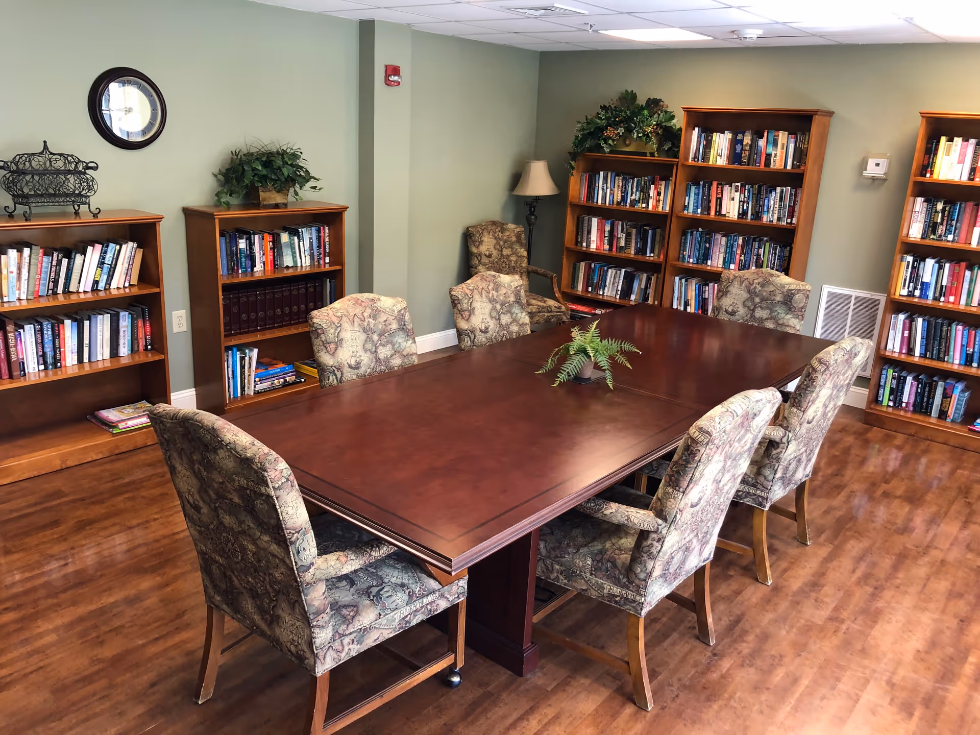 Library-style common room with a large wooden table, upholstered chairs, and bookshelves against green walls.
