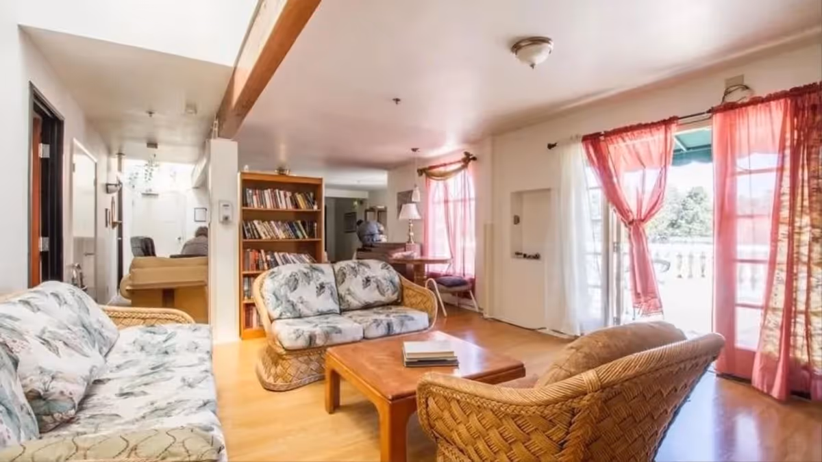 A cozy living room area with wicker furniture including a sofa, loveseat, and armchair with floral cushions, a wooden coffee table with books on it, a bookshelf filled with books, and large windows with sheer white and pink curtains allowing natural light to fill the room.