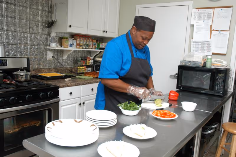 A chef wearing a blue shirt, black apron, and black hat is preparing food in a kitchen. He is slicing a cucumber on a cutting board placed on a stainless steel counter. There are plates with sliced tomatoes, leafy greens, and other food items on the counter. The kitchen has white cabinets, a stove with a pot, a microwave, and a bulletin board with papers pinned on it.