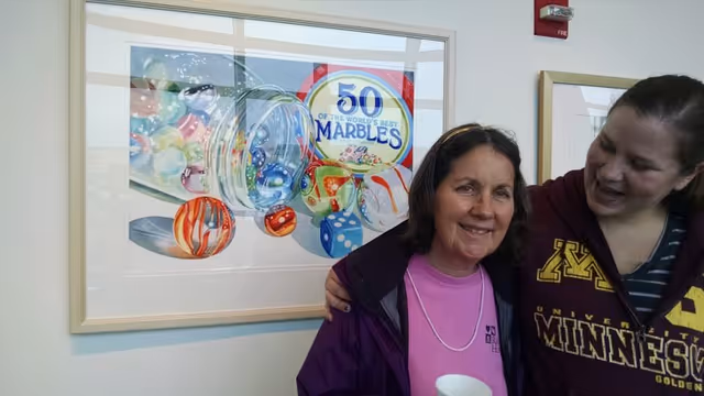 Two women smiling and standing close together in front of a framed picture of colorful marbles and a container labeled '50 of the World's Best Marbles'. One woman is wearing a purple jacket and pink shirt, the other is wearing a maroon University of Minnesota sweatshirt.