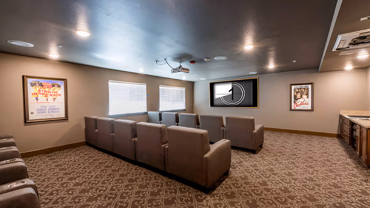 A cozy movie theater room with two rows of comfortable armchairs facing a large screen displaying a countdown timer. The room has patterned carpet, beige walls, two windows with blinds, and framed classic movie posters on the walls. There is also a kitchenette area with cabinets and a countertop on the right side.
