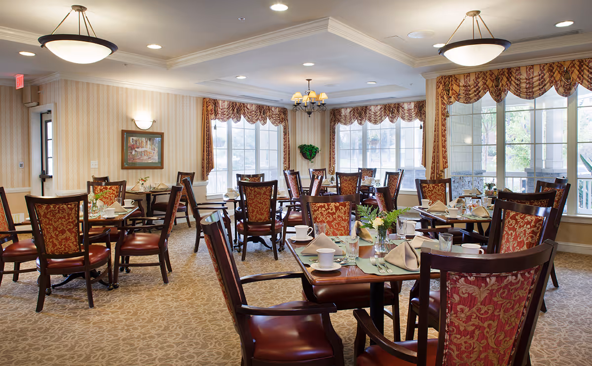 A well-lit dining room with multiple wooden tables and chairs featuring red and gold patterned upholstery. Each table is set with cups, glasses, napkins, and small floral centerpieces. Large windows with decorative valances allow natural light to fill the room, and there are ceiling lights and chandeliers providing additional lighting. The walls have a striped wallpaper and framed artwork.