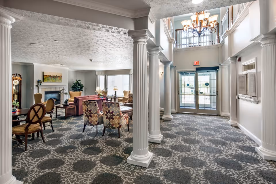 Spacious senior living facility lobby with tall white columns, patterned carpet, multiple seating areas, a fireplace, and glass entry doors.